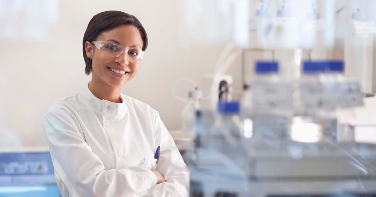 A woman in a white lab coat stands and smiles in her lab