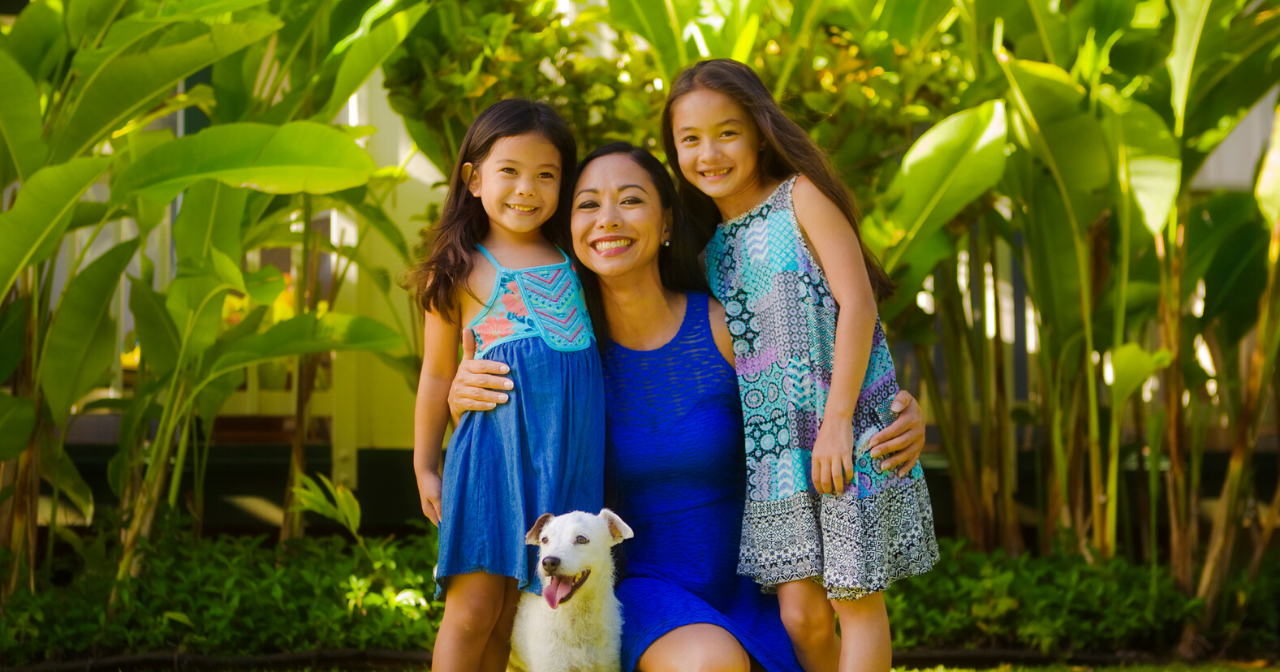 A mother, her two daughters, and a dog smile in a tropical setting.