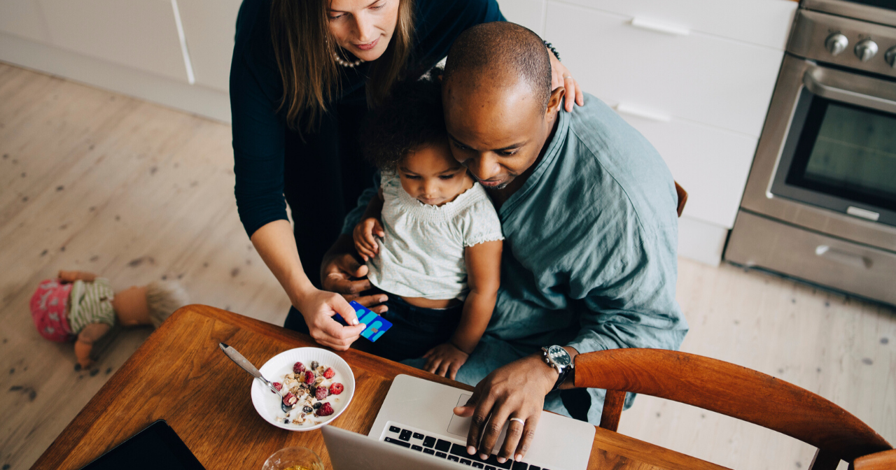 A father sits with his child on his lap at the breakfast table while the child's mother leans over his shoulder to look at his laptop screen. 