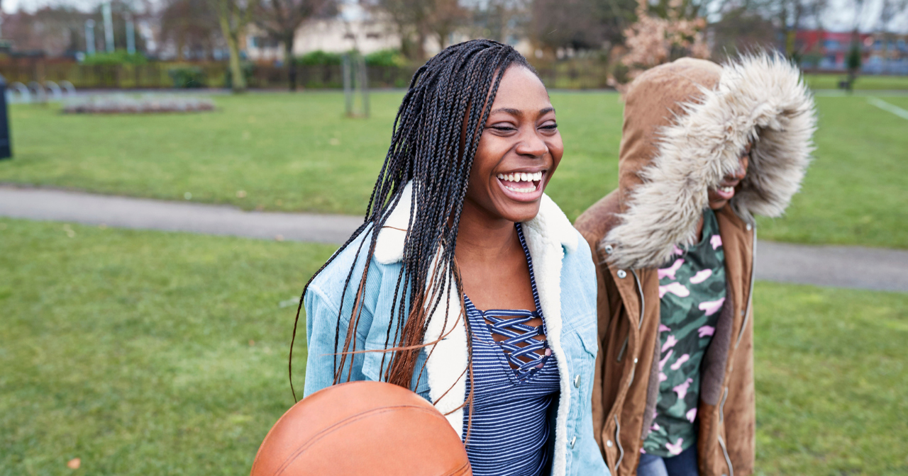 Two Black girls laugh while walking through a park.