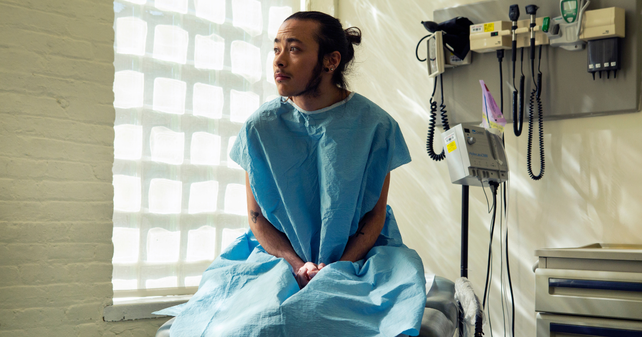 A genderqueer person sits in an exam room wearing a hospital gown. 