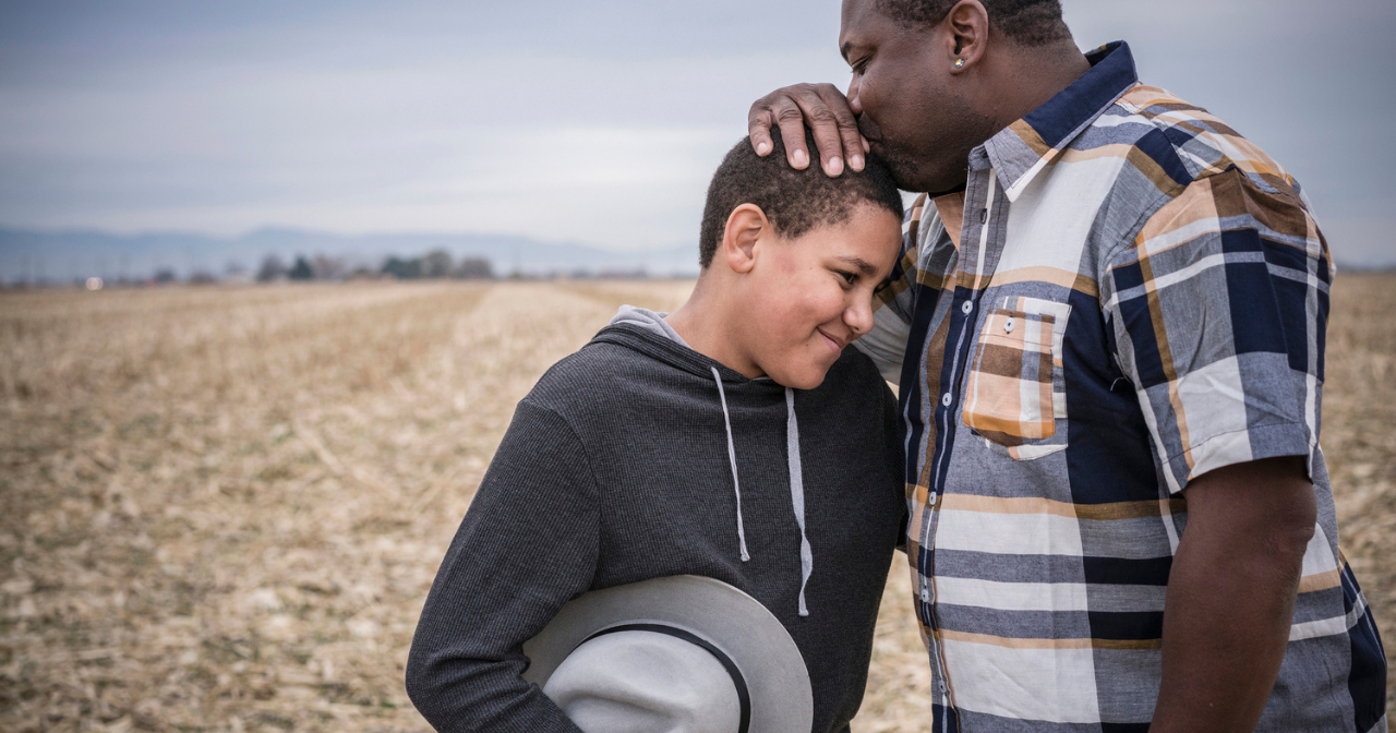 A father and son stand a in a field and the father kisses the top of his son's head. 