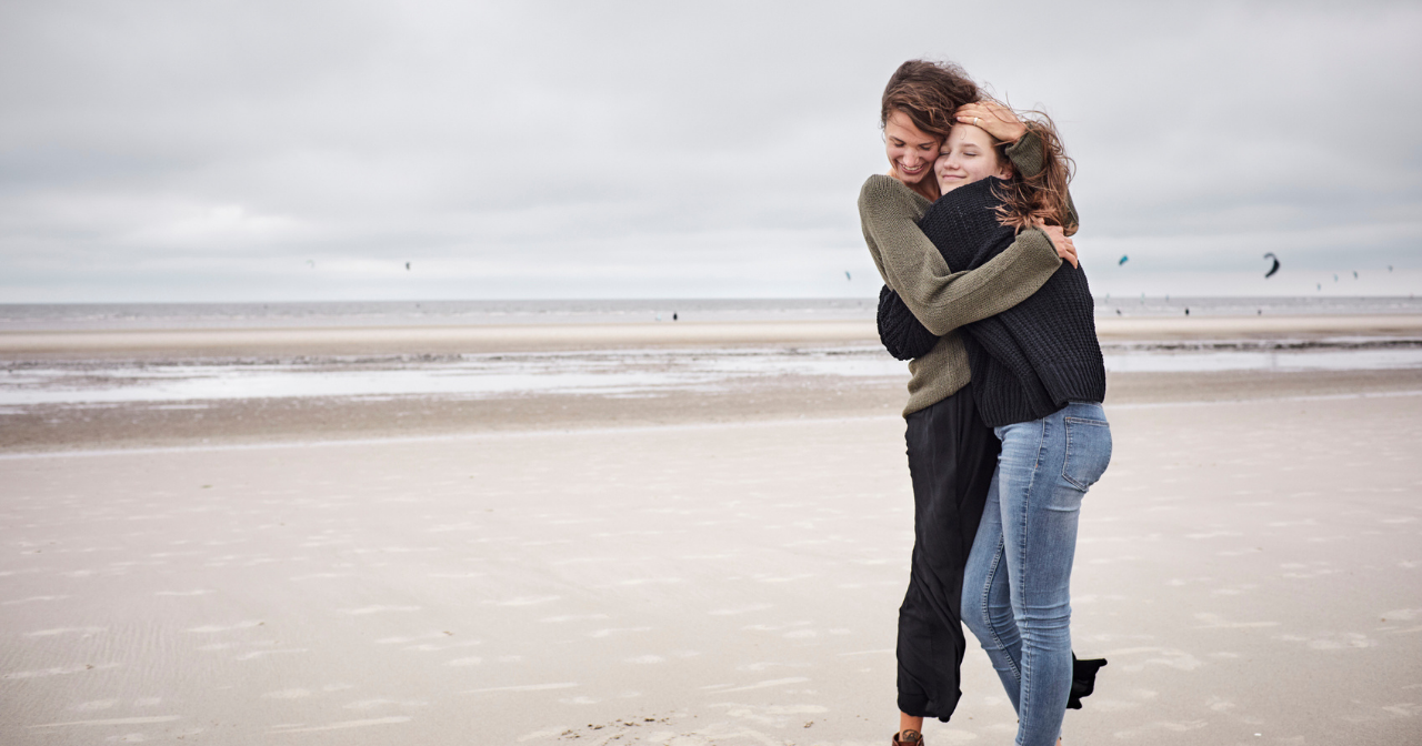 A mother and daughter laugh and hug while standing on a beach. 