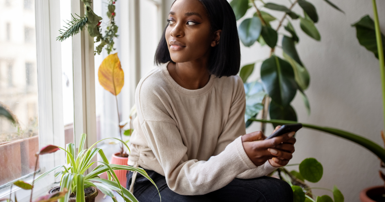A Black woman sits next to a window and looks out contemplatively. 