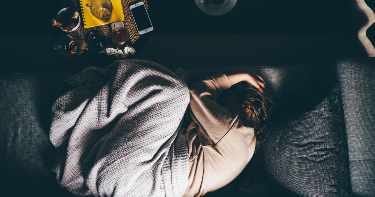 A woman lays curled up under a blanket on a sofa while holding her head and overwhelmed. 