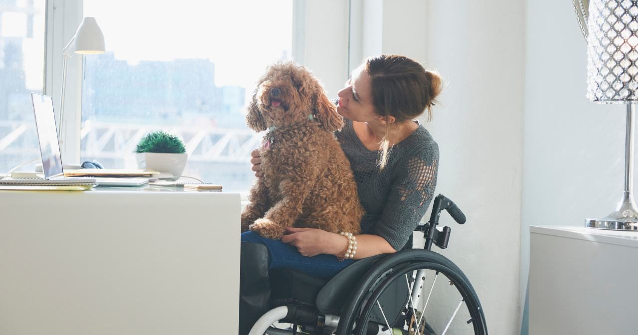 A woman in a wheelchair sits at her desk with her dog in her lap. 