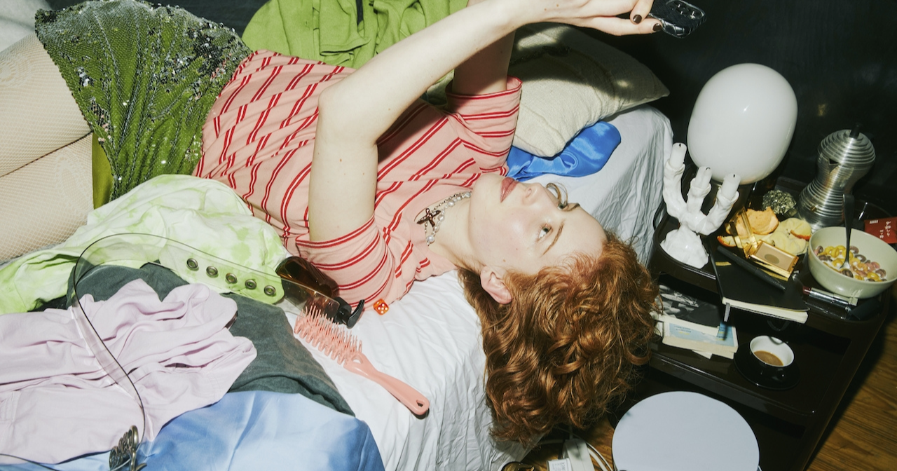 A young woman scrolls on her phone while laying on a messy bed. 