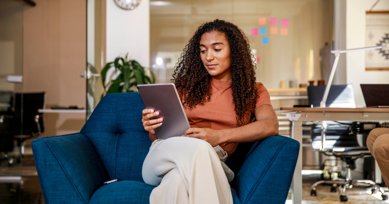 A woman sits in a chair and reads on a tablet.