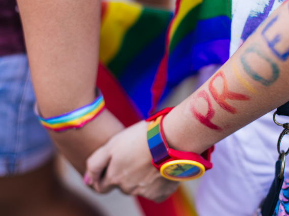 To people holding hands with rainbow accessories and the word PRIDE written on one forearm
