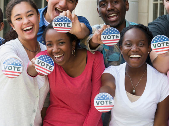 A group of teens smile and hold out red, white, and blue buttons that read, "Vote."