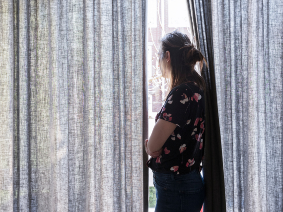 An image of a woman hugging herself and looking out of a window mostly covered by curtains. 