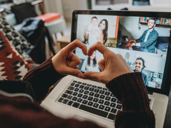 A person makes a heart with their fingers in front of their computer during a Zoom call with friends. 