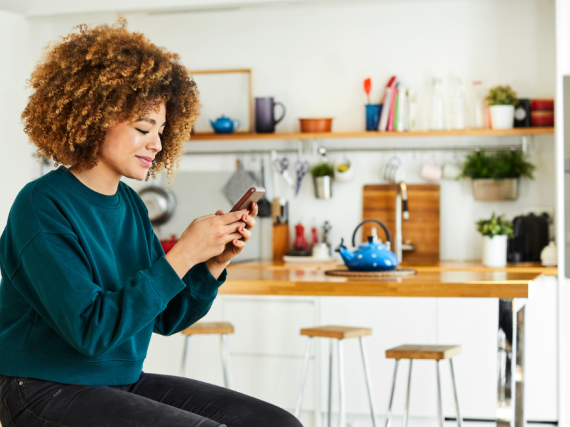 A woman sits on a stool in the kitchen with her phone in her hands. 