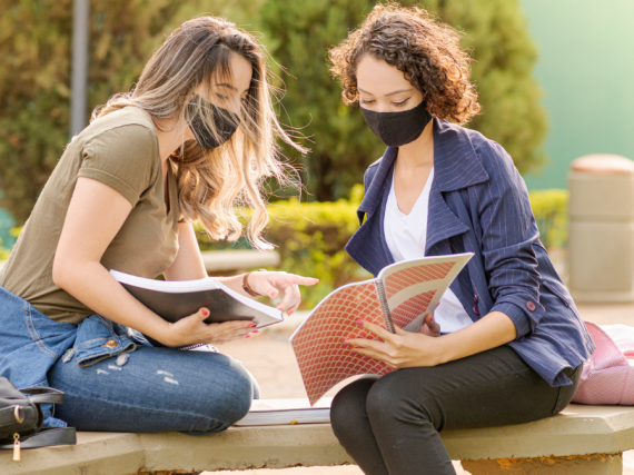 Two women wearing masks sit on a bench outside and study together. 