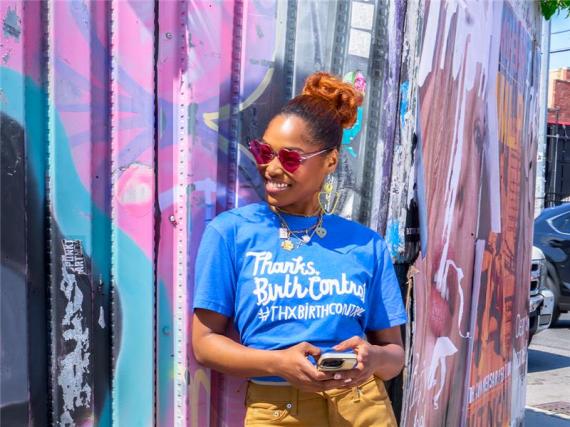 A woman in a shirt that says #ThxBirthControl smiles while leaning against a wall with a mural on it. 