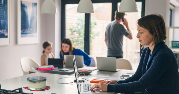 A family of four sits in the kitchen and works on their computers and phones. 
